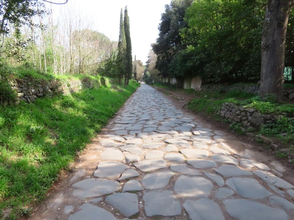 Original Roman-era paving stretching into the distance between grassy banks - stone wall on the left and trees on the right.