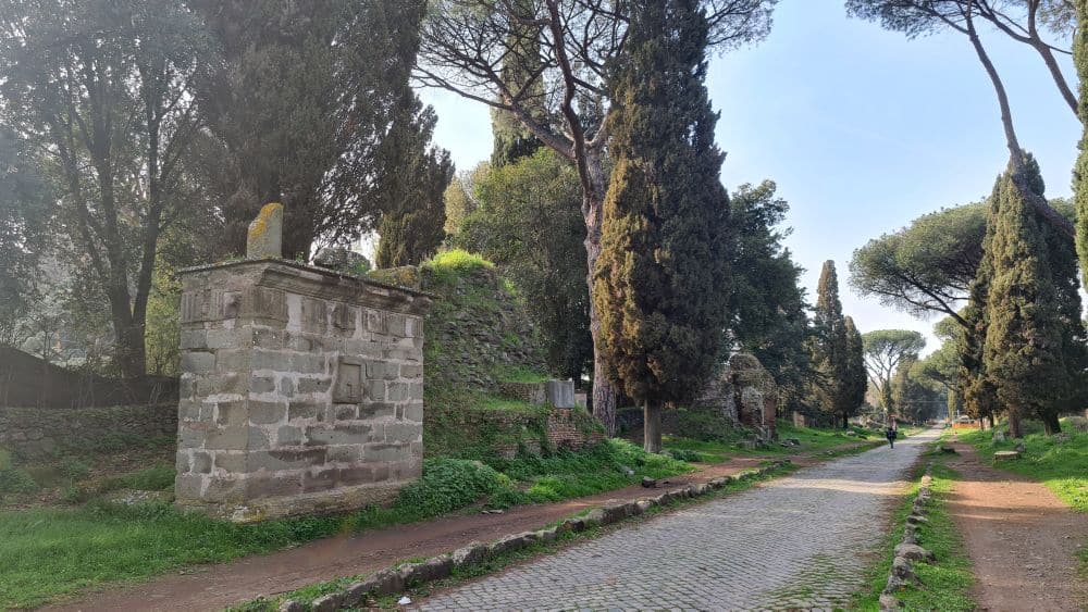 The road, with newer paving, and funerary monuments in a row along it. The first is a piece of wall, in good shape. The second is a pile of stones, overgrown with grass,  and the third is, like the first, visibly a monument.