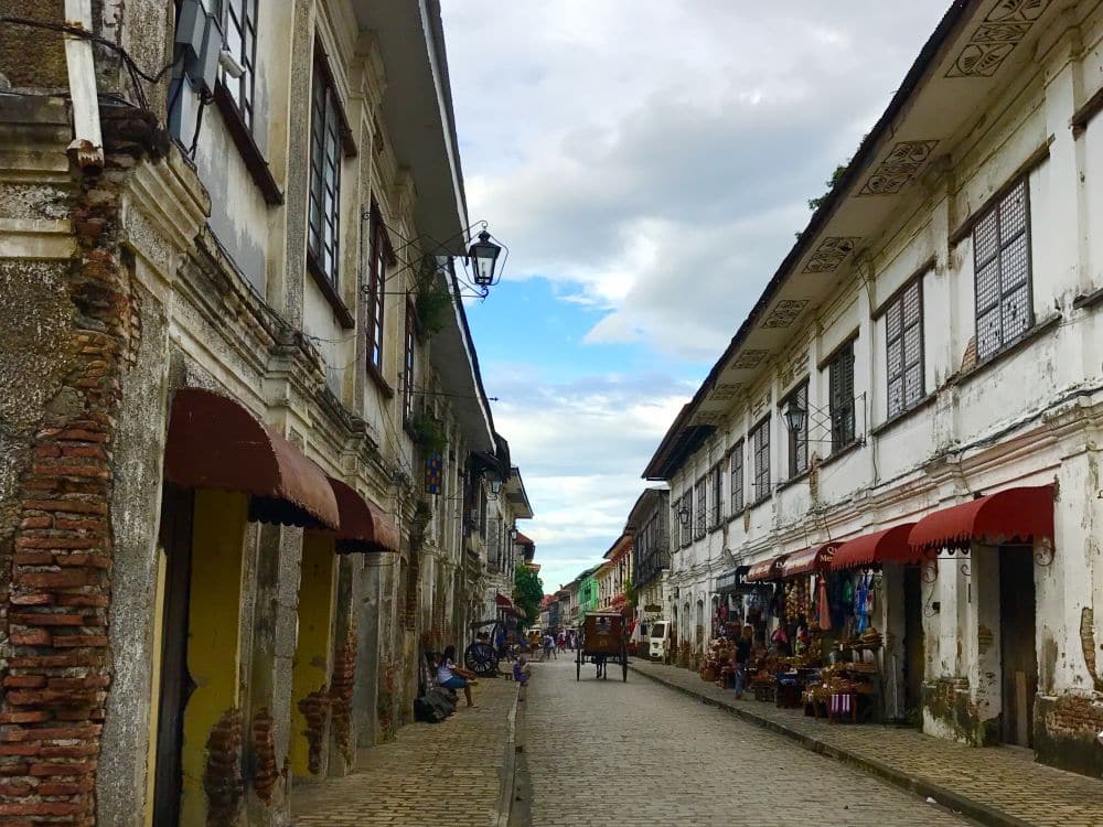 A view bown a cobbled street: 2-story rows on both sides, with shops on the ground floors. Rather poor condition.