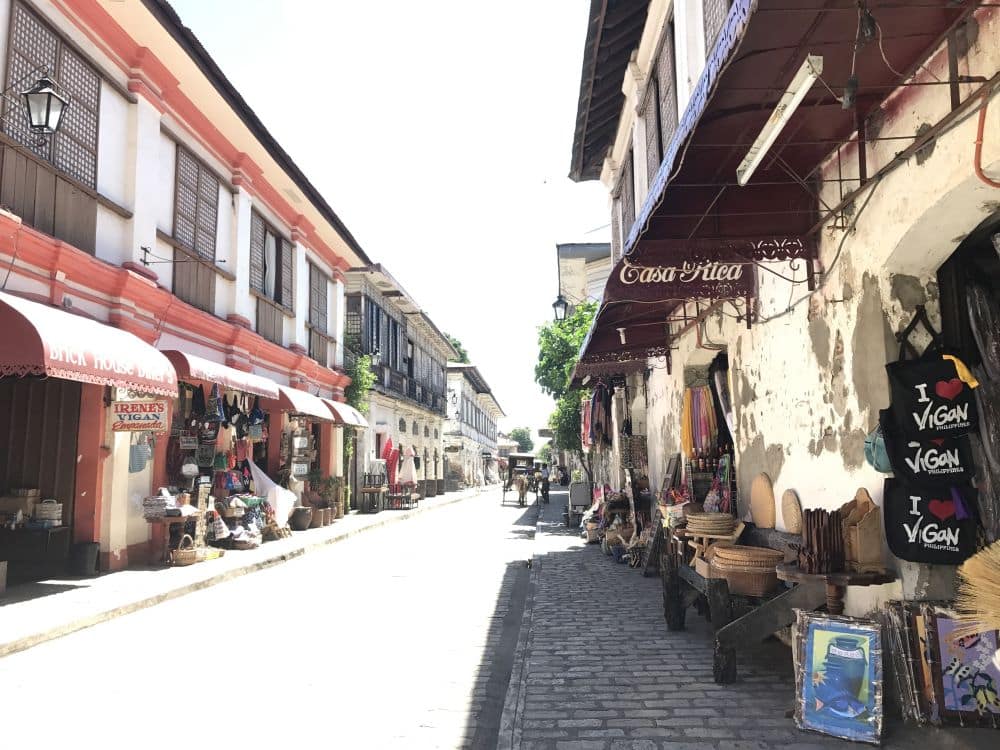 City street with 2-story rows on either side and projecting shades over the sidewalks. Lots of souvenir shops.