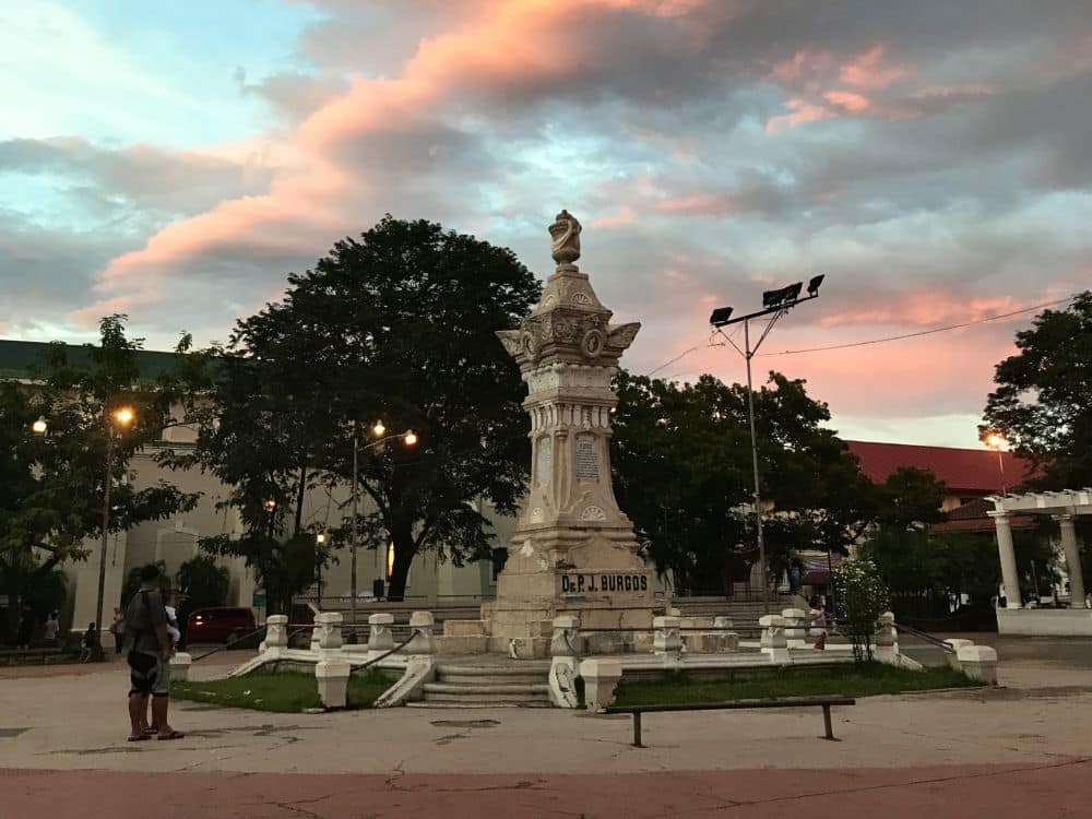 A monument in a plaza with a sunset sky above the far buildings.