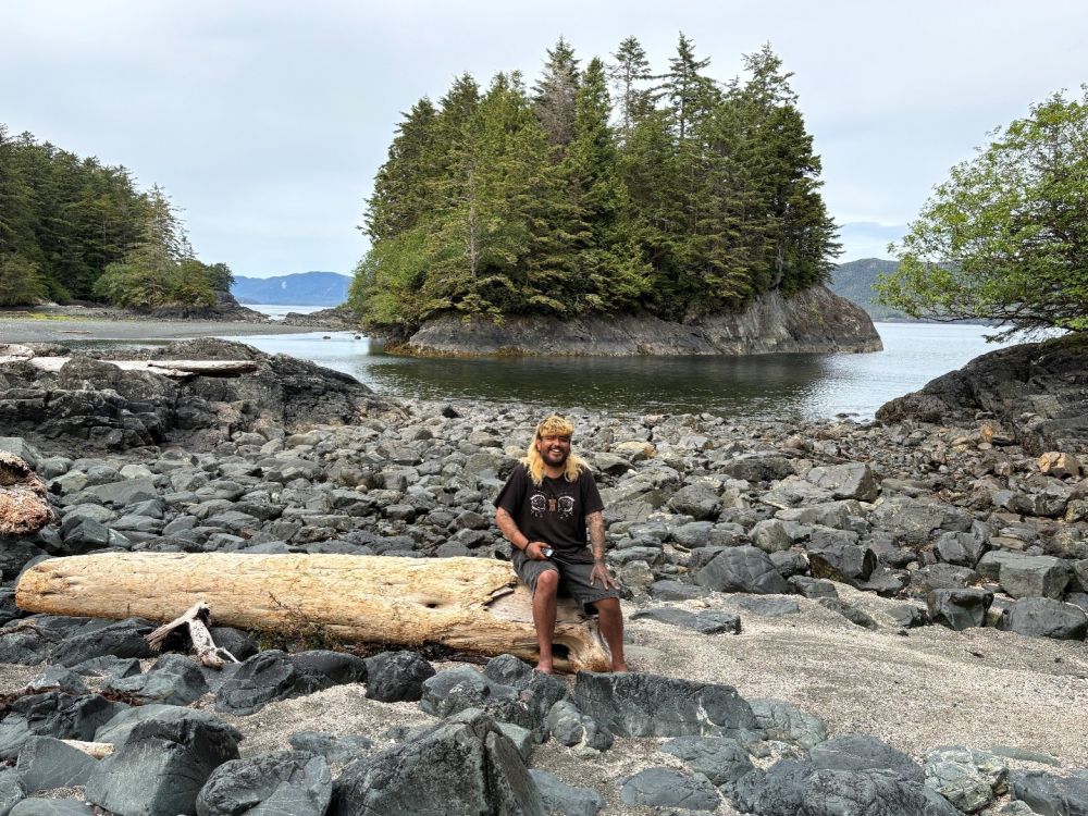 A young man sits on a fallen tree trunk on a rocky shore with a small wooded island behind him.