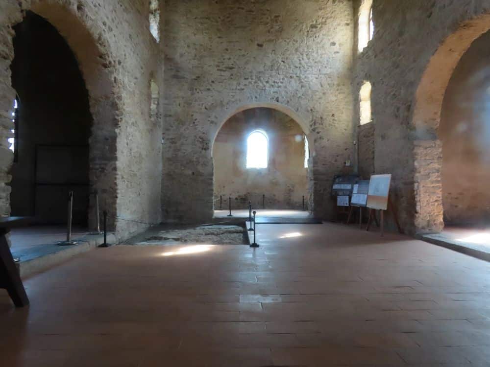 A large empty room, an apse at the far end and an larger apse on both sides. One section of the floor is not tiled and has a cordon around it.