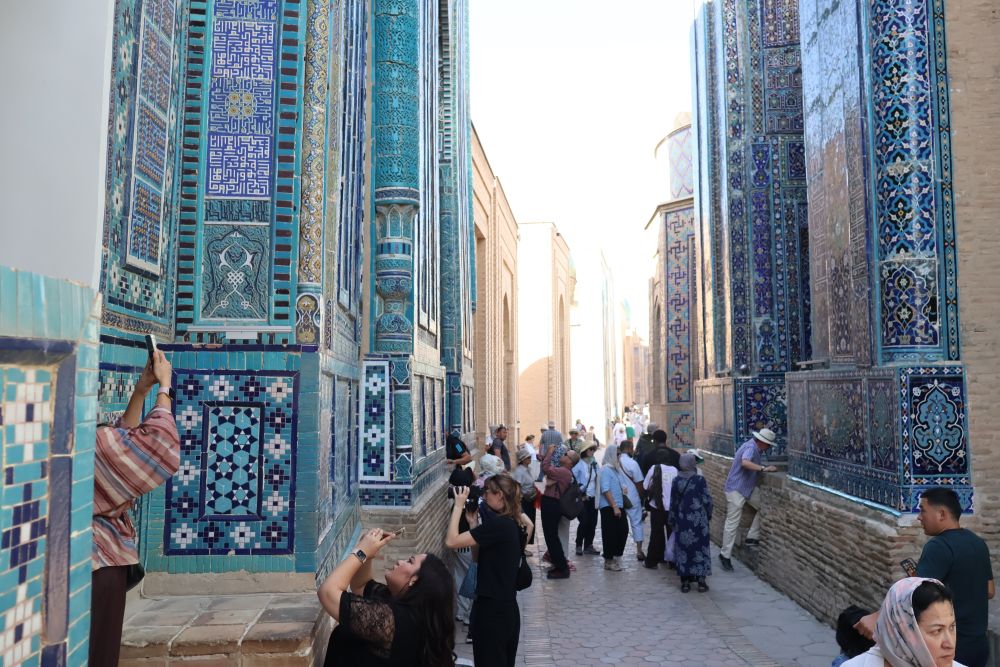 Very tall colorful buildings line both sides of a narrow lane, with many tourists trying to take pictures.
