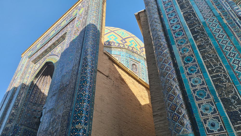 Looking up at two mausoleums: the fronts are covered in tile, mostly blue, and the dome of one is partly visible, also mostly blue.